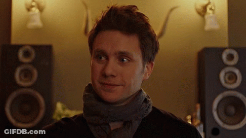 A Man With A Scarf Around His Neck Is Smiling In Front Of A Pair Of Speakers GIF