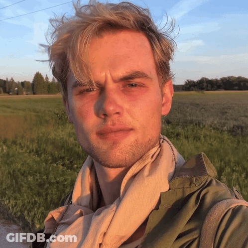 A Man With A Scarf Around His Neck Stands In A Field GIF