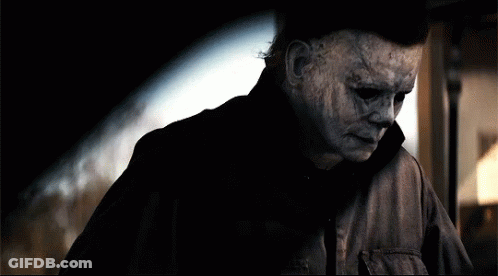 A Man With A Scary Mask On His Face Is Standing In A Dark Room . GIF
