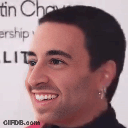 A Man With A Shaved Head And Earrings Is Smiling In Front Of A Sign That Says Tin Chow . GIF