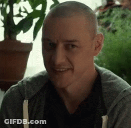 A Man With A Shaved Head Is Smiling And Looking At The Camera While Sitting In Front Of A Potted Plant . GIF