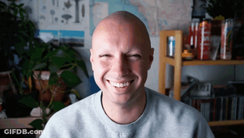 A Man With A Shaved Head Is Smiling In Front Of A Plant GIF