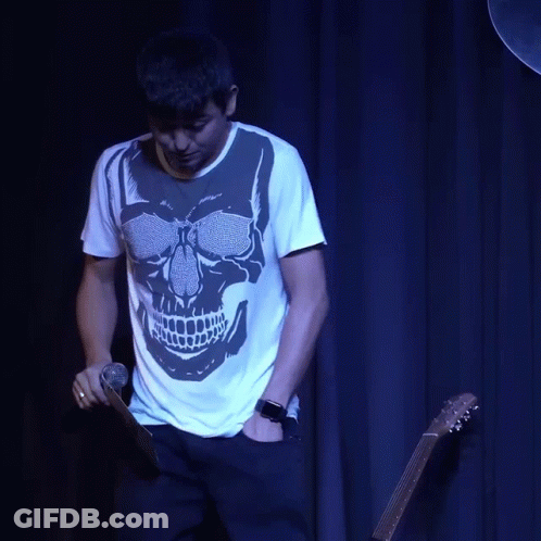A Man With A Skull On His Shirt Is Holding A Guitar And Talking On A Cell Phone GIF