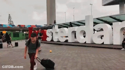 A Man With A Suitcase Is Walking In Front Of A Large Amsterdam Sign GIF