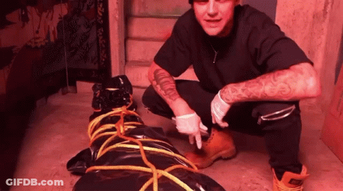 A Man With A Tattoo On His Arm Is Kneeling Next To A Black Bag Tied With Rope GIF