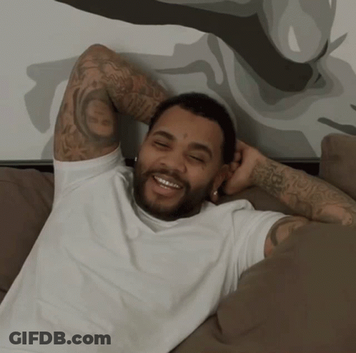 A Man With A Tattoo On His Arm Is Laying On A Couch And Smiling GIF