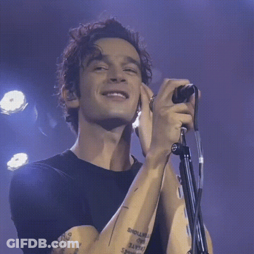 A Man With A Tattoo On His Arm Is Singing Into A Microphone While Smiling GIF