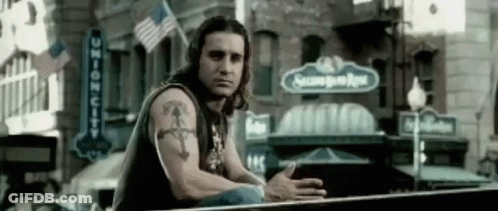 A Man With A Tattoo On His Arm Is Sitting On A Railing In Front Of A Building With A Sign That Says Second Base GIF