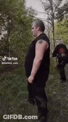 A Man With A Tattoo On His Arm Is Standing In The Woods Next To A Woman . GIF