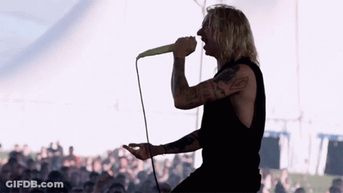 A Man With A Tattoo On His Arm Singing Into A Microphone With A Crowd Behind Him GIF
