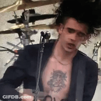 A Man With A Tattoo On His Chest Is Playing A Guitar In Front Of A Microphone GIF