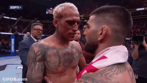 A Man With A Tattoo On His Chest Stands Next To Another Man With A Bud Light Banner Behind Him GIF
