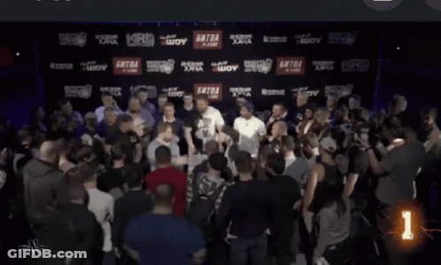 A Man With A Tattoo On His Face Stands In A Crowd With A Security Guard Behind Him GIF
