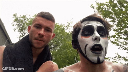 A Man With A Towel Around His Neck Is Standing Next To A Man With A Scary Face Painted On His Face GIF