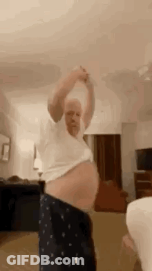 A Man With A Very Large Belly Is Dancing In A Living Room With A Woman . GIF
