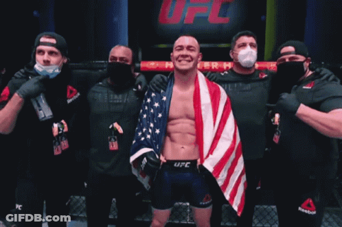 A Man With An American Flag Wrapped Around His Shoulders Stands In Front Of A Sign That Says Fc GIF