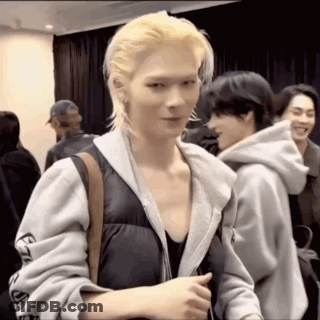 A Man With Blonde Hair And A Hoodie Is Standing In A Room With Other People . GIF