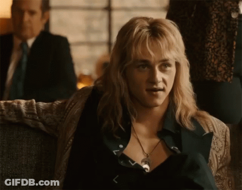A Man With Blonde Hair And A Necklace Is Sitting On A Couch In Front Of A Man In A Suit . GIF