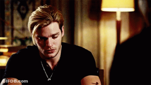 A Man With Blonde Hair And A Tattoo On His Arm Is Sitting At A Table With A Lamp In The Background . GIF