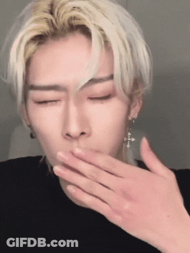 A Man With Blonde Hair And Earrings Is Covering His Mouth With His Hand GIF