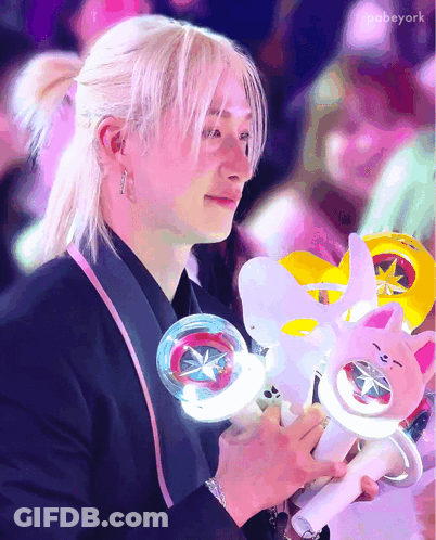 A Man With Blonde Hair Is Holding A Light Stick And A Stuffed Animal GIF