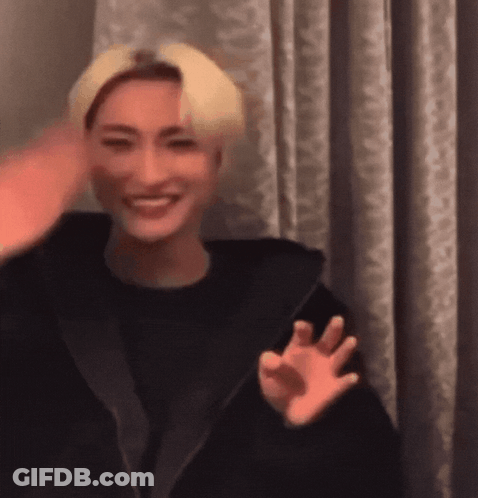 A Man With Blonde Hair Is Smiling And Waving At The Camera While Wearing A Black Jacket . GIF