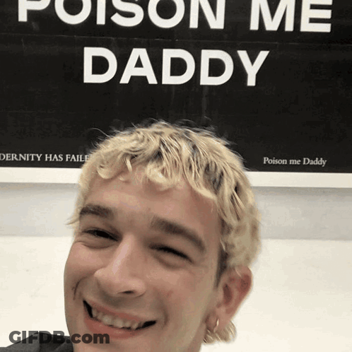A Man With Blonde Hair Is Smiling In Front Of A Sign That Says Poison Me Daddy GIF