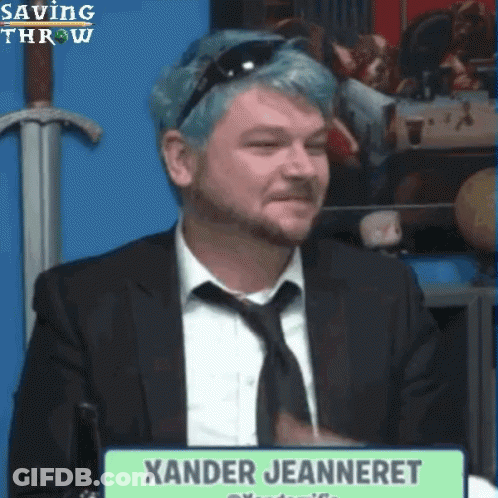 A Man With Blue Hair Is Sitting In Front Of A Sign That Says Xander Jeanneret GIF