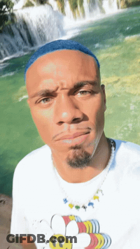 A Man With Blue Hair Is Wearing A White Shirt With A Bee On It In Front Of A Waterfall GIF