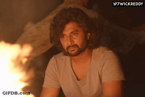 A Man With Curly Hair And A Beard Is Sitting In Front Of A Fire With The Hashtag 7Wickreddy Above Him GIF
