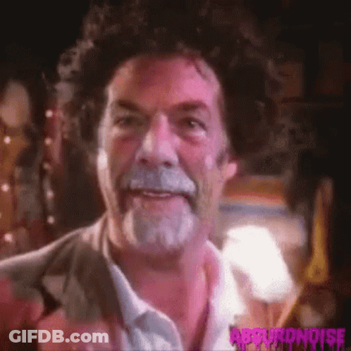 A Man With Curly Hair And A Beard Is Smiling And Looking At The Camera . GIF