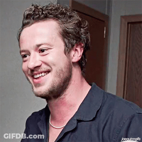 A Man With Curly Hair And A Beard Is Smiling And Wearing A Necklace . GIF