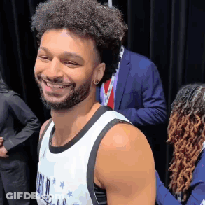 A Man With Curly Hair And A Beard Is Smiling For The Camera While Wearing A Basketball Jersey . GIF