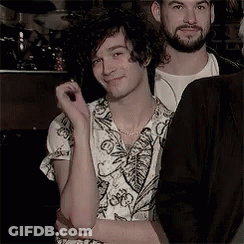 A Man With Curly Hair And A Beard Is Standing Next To Another Man In A Room . GIF