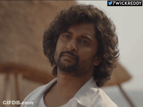 A Man With Curly Hair And A Beard Looks At The Camera With A 7Wickreddy Logo Behind Him GIF