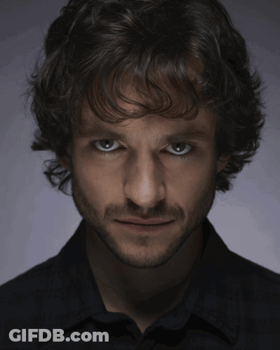 A Man With Curly Hair And A Beard Looks At The Camera With A Serious Look On His Face GIF