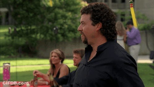 A Man With Curly Hair And A Beard Stands In Front Of A Table GIF