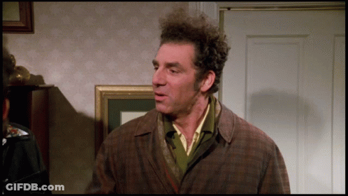 A Man With Curly Hair And A Plaid Jacket Stands In Front Of A Door GIF