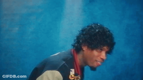 A Man With Curly Hair And A Varsity Jacket Is Standing In Front Of A Blue Wall . GIF