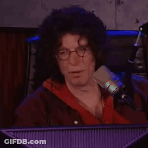 A Man With Curly Hair And Glasses Is Sitting In Front Of A Microphone In Front Of A Computer Monitor . GIF