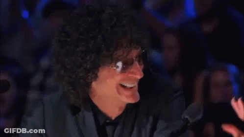 A Man With Curly Hair And Sunglasses Is Smiling In Front Of A Crowd In A Dark Room . GIF