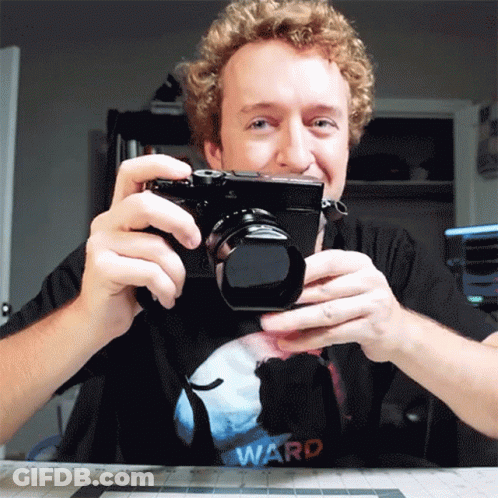 A Man With Curly Hair Is Holding A Camera And Wearing A Ward Shirt GIF