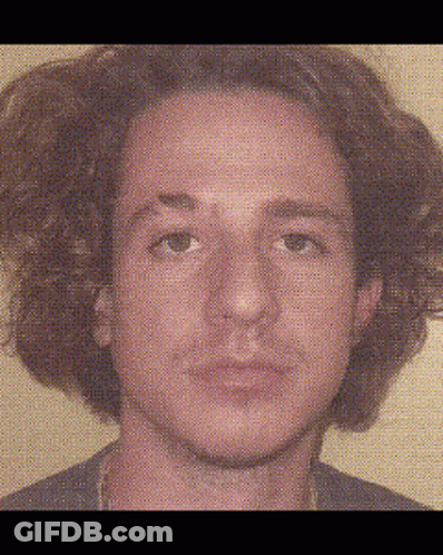 A Man With Curly Hair Is Looking At The Camera With A Blurred Background Behind Him . GIF