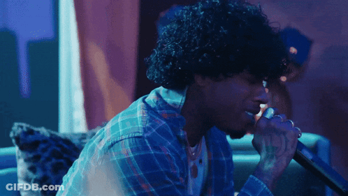 A Man With Curly Hair Is Singing Into A Microphone In A Dark Room GIF