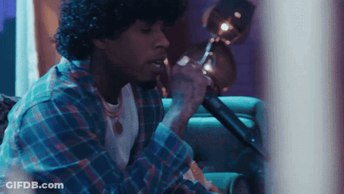 A Man With Curly Hair Is Singing Into A Microphone While Sitting On A Couch GIF