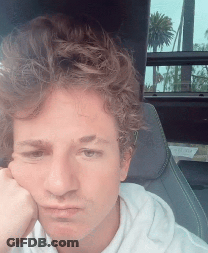 A Man With Curly Hair Is Sitting In A Car With His Hand To His Face GIF