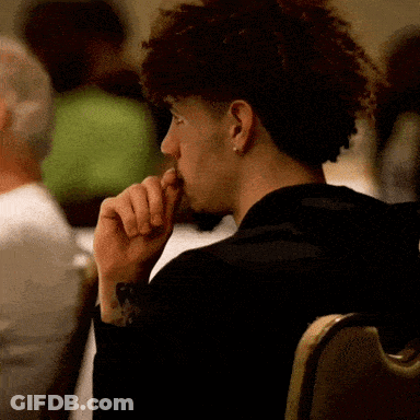A Man With Curly Hair Is Sitting In A Chair And Looking At Something GIF