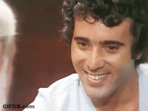 A Man With Curly Hair Is Smiling And Talking To Another Man . GIF