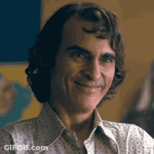 A Man With Curly Hair Is Smiling For The Camera While Wearing A White Shirt . GIF