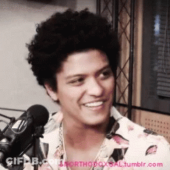A Man With Curly Hair Is Smiling In Front Of A Microphone GIF
